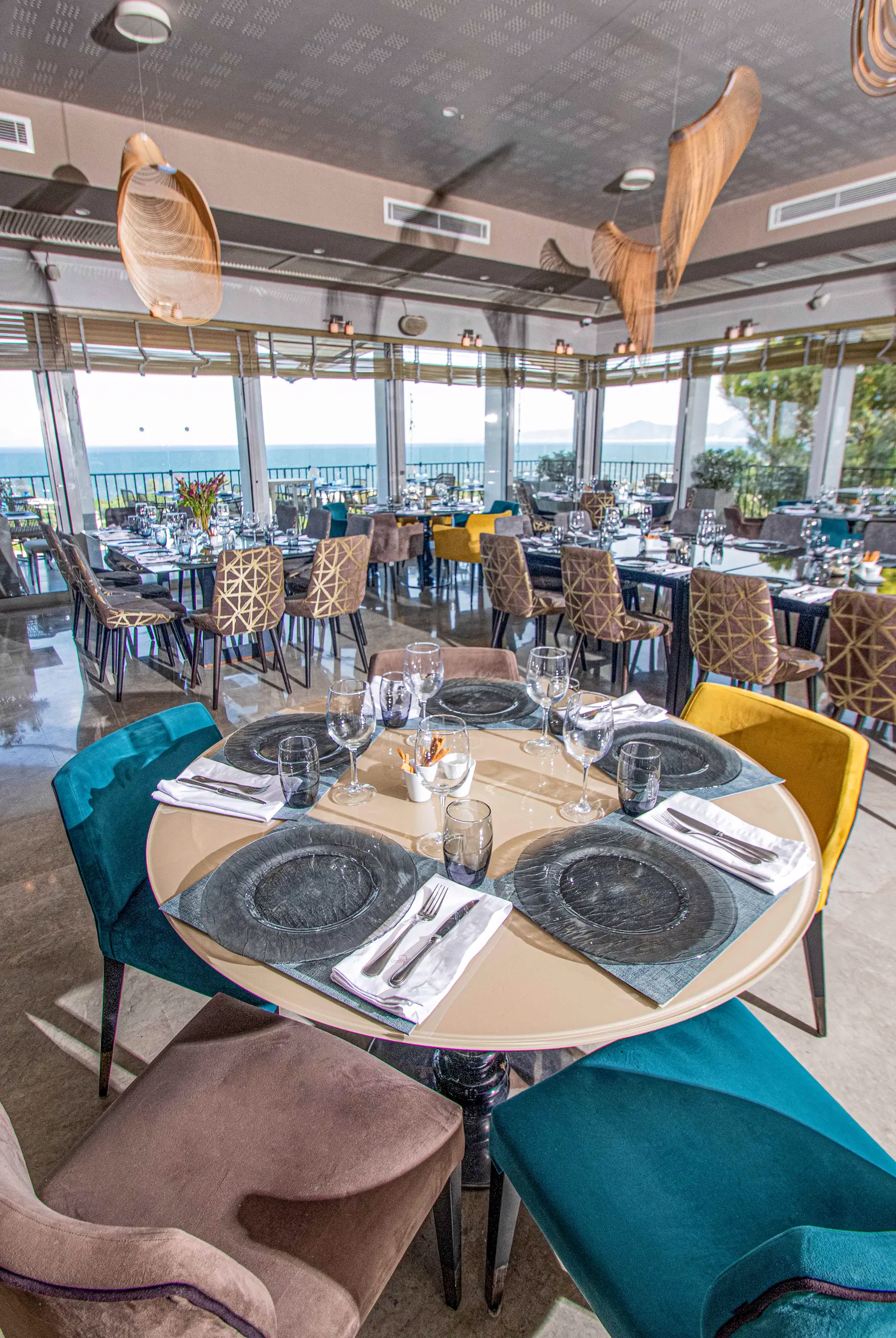 Le Restô's panoramic dining room in full daylight: teal and gold velvet armchairs, floor-to-ceiling windows open to the sea at Carthage
