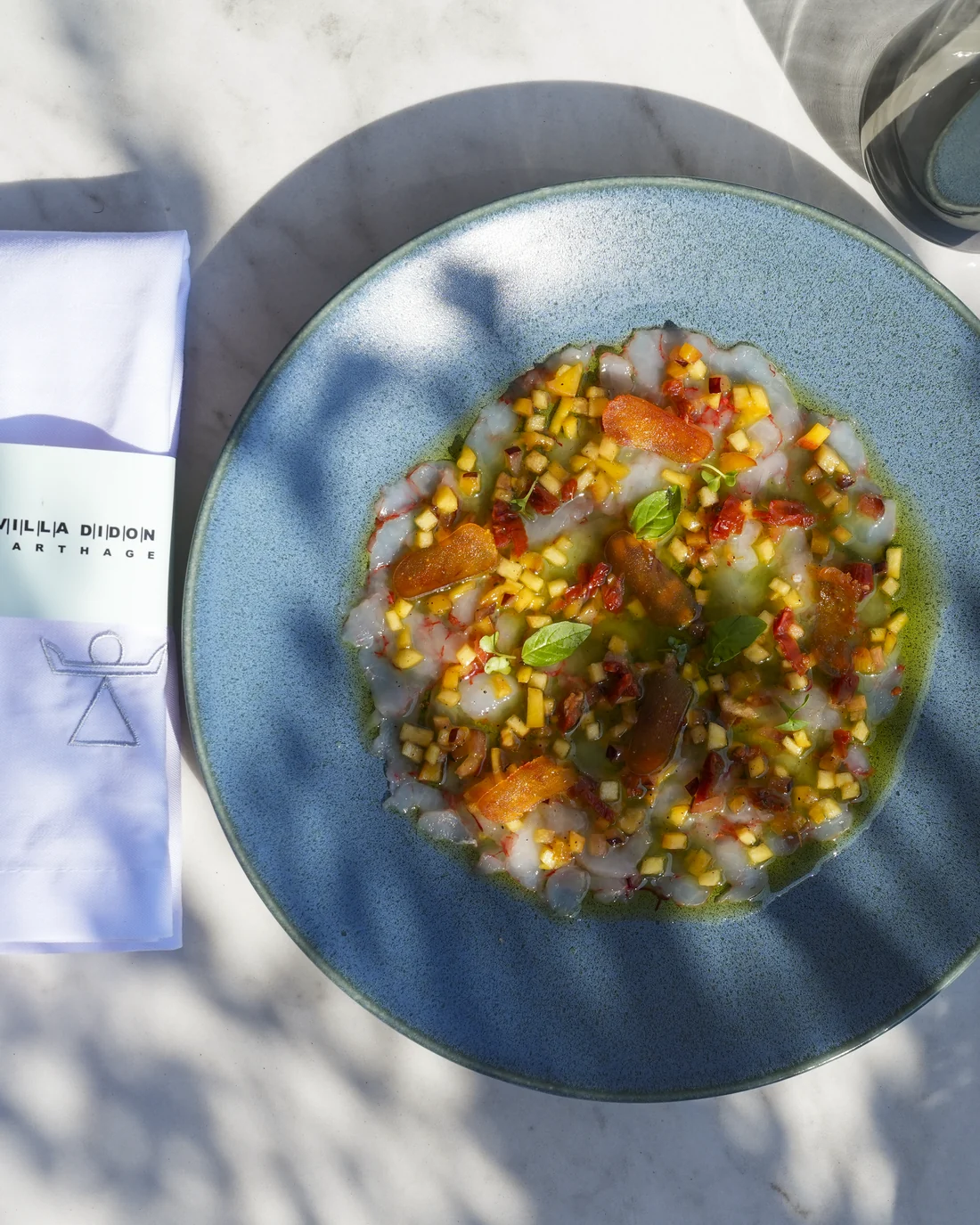 Sea bream ceviche from above: raw fish, a brunoise of citrus and peppers, basil, in a turquoise plate on marble, Villa Didon napkin and shadows of foliage