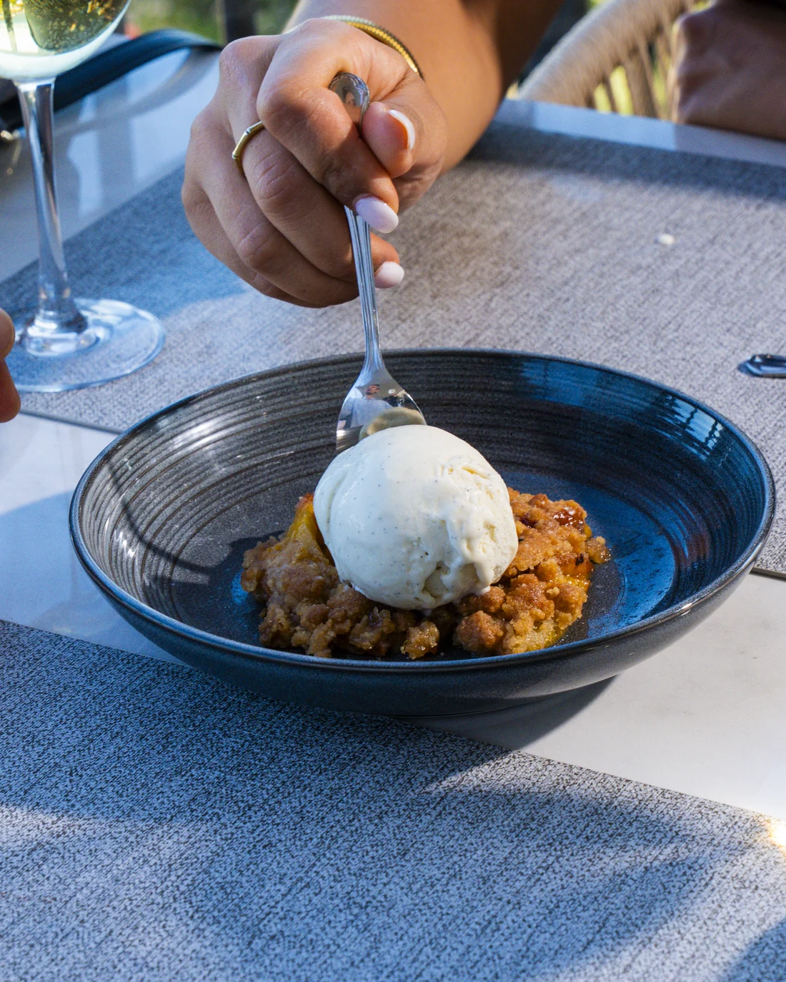 Dessert served with a spoon: crumble and a quenelle of ice cream in a blue bowl, a glass of white wine behind