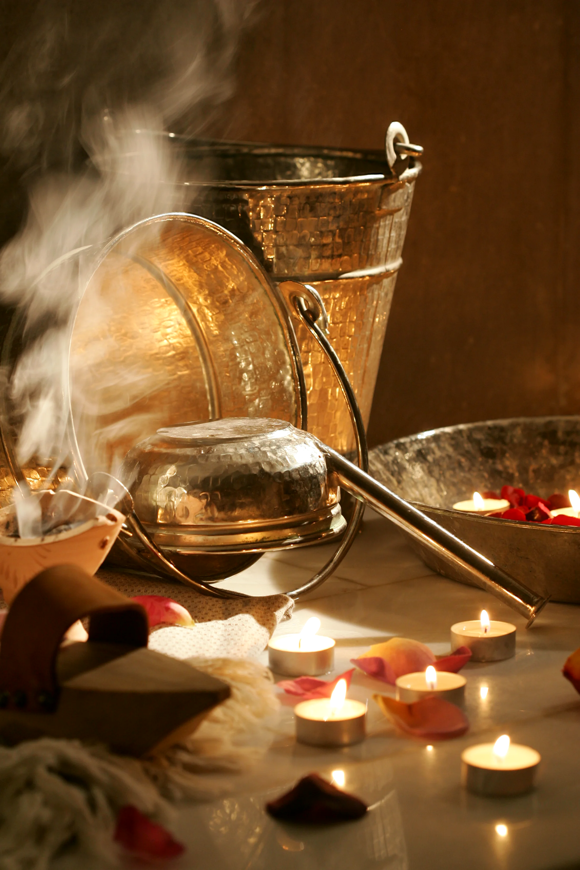 A detail of the hammam ritual: copper pails and bowls, steam rising through candlelight, rose petals